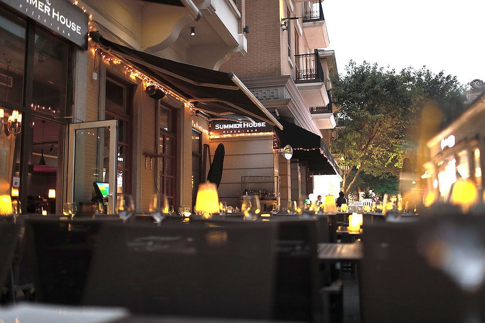 Outdoor dining area at dusk, tables with candles glowing, under "SUMMER HOUSE Pizza Bar" sign. Warm ambiance, trees in the background.