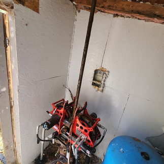 A red and black hydraulic well pump puller in operation inside a small shed, lifting a long, rusted vertical pipe upwards toward the ceiling, situated next to a blue pressure tank.
