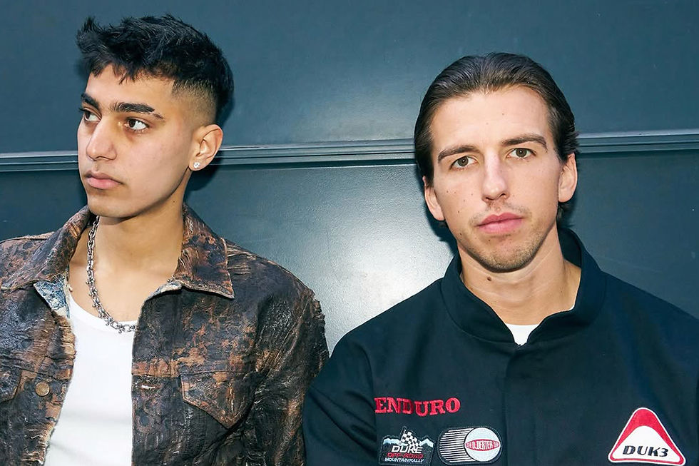 Omar+ & Josh Baker against a blue wall. One is wearing a brown jacket, the other dark clothing with patches, both with serious expressions.