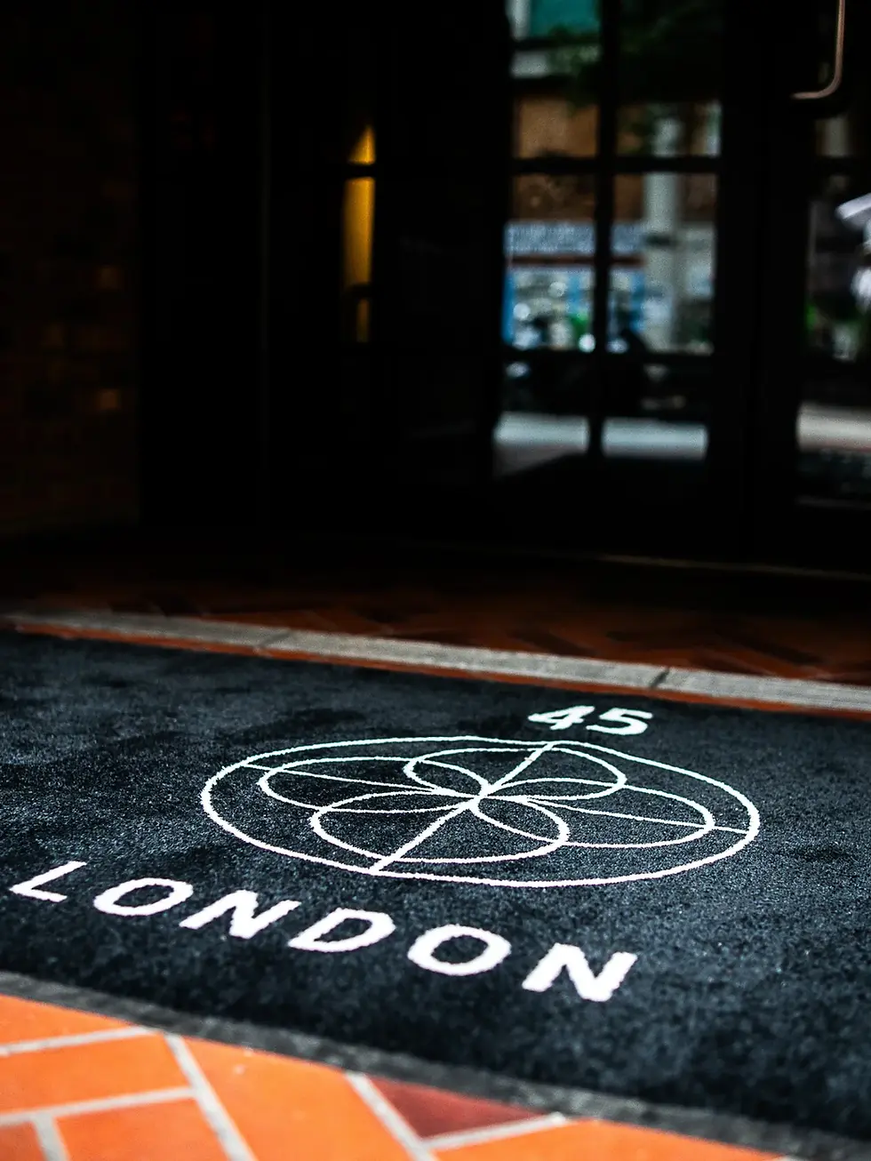 Dark entrance with a black mat featuring a white circular design and "45 LONDON" text. Blurry glass door in the background. 45 London on 45 Curtain Road. 