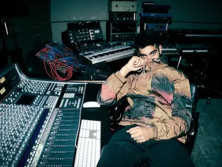 OMAR+ in a camouflage jacket, sitting at a mixing desk in the studio, holding his collar. Equipment and a keyboard are visible in the background.