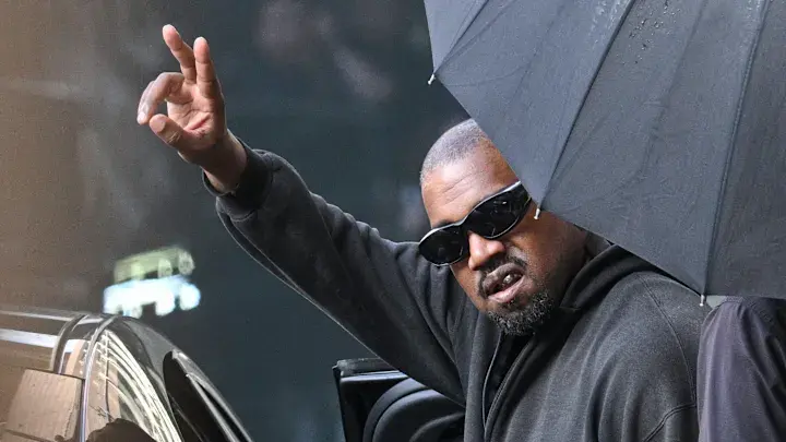 Kanye West (Ye) wearing glasses, raising his hand while sitting in a car under an umbrella. Grey outdoor background, dynamic atmosphere.