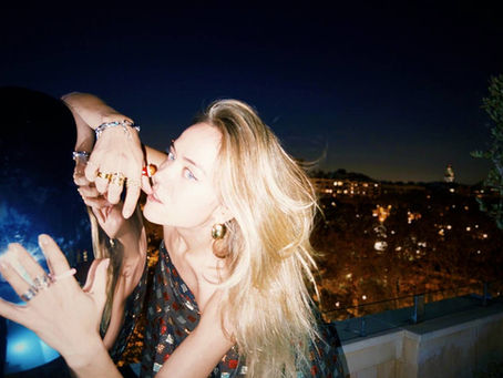 Elizabeth Lip with long blonde hair against a night-time city backdrop. She is wearing a sparkling dress and jewellery, looking into the camera with a thoughtful expression.
