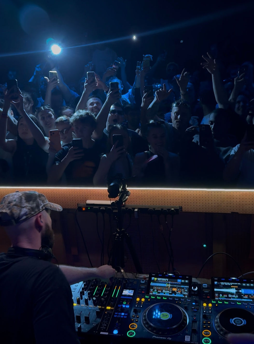 NOTION mixing tracks on a console in a dimly lit club during Mixmag Cover Moment. A lively crowd in the background eagerly takes photos with phones.
