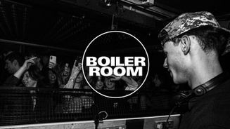 A DJ wearing headphones at a Boiler Room party. A crowd surrounds him, with many people filming on their phones. Energetic and lively mood. Black-and-white photo.