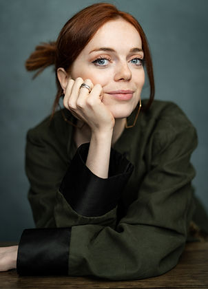 Portrait of El Blackwood. El is wearing a deep green jacket with black cuffs, and resting her face on her hand.