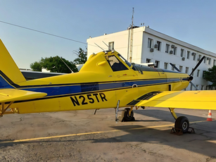 The image shows an Air Tractor 502A, a bright yellow and blue single-engine agricultural aircraft, parked on the tarmac in front of a white airport or aviation facility building. The aircraft bears the registration number N25TR, which is prominently displayed on the fuselage.