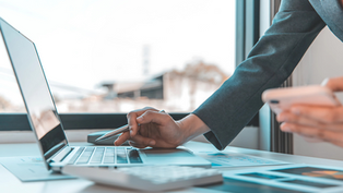A professional woman multitasks with a laptop and smartphone, surrounded by financial documents and a calculator. Perfect for topics on business analysis, digital tools, remote work, and productivity in a modern office setting.