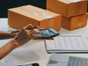 A person using a calculator and checking shipping data beside cardboard boxes and a laptop, showing logistics and delivery cost calculation.