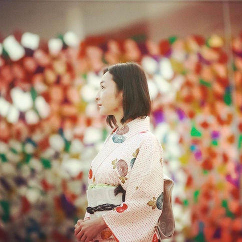 Early autumn morning in Okazaki Park, Kyoto, with soft pre-sunrise light illuminating a beautiful person. The scene includes a temporary colorful leaf-like glass backdrop and the giant Torii gate in the background.