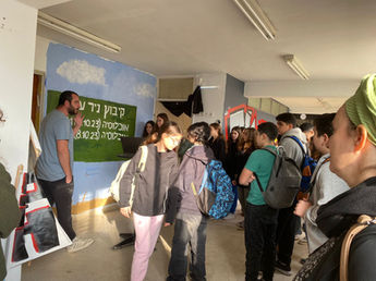 קבוצת חניכים בסיור בתערוכה ומדריך שמסביר על ציור בו כתוב מספר התושבים בקיבוץ ניר עוז לפני ואחרי השביעי באוקטובר