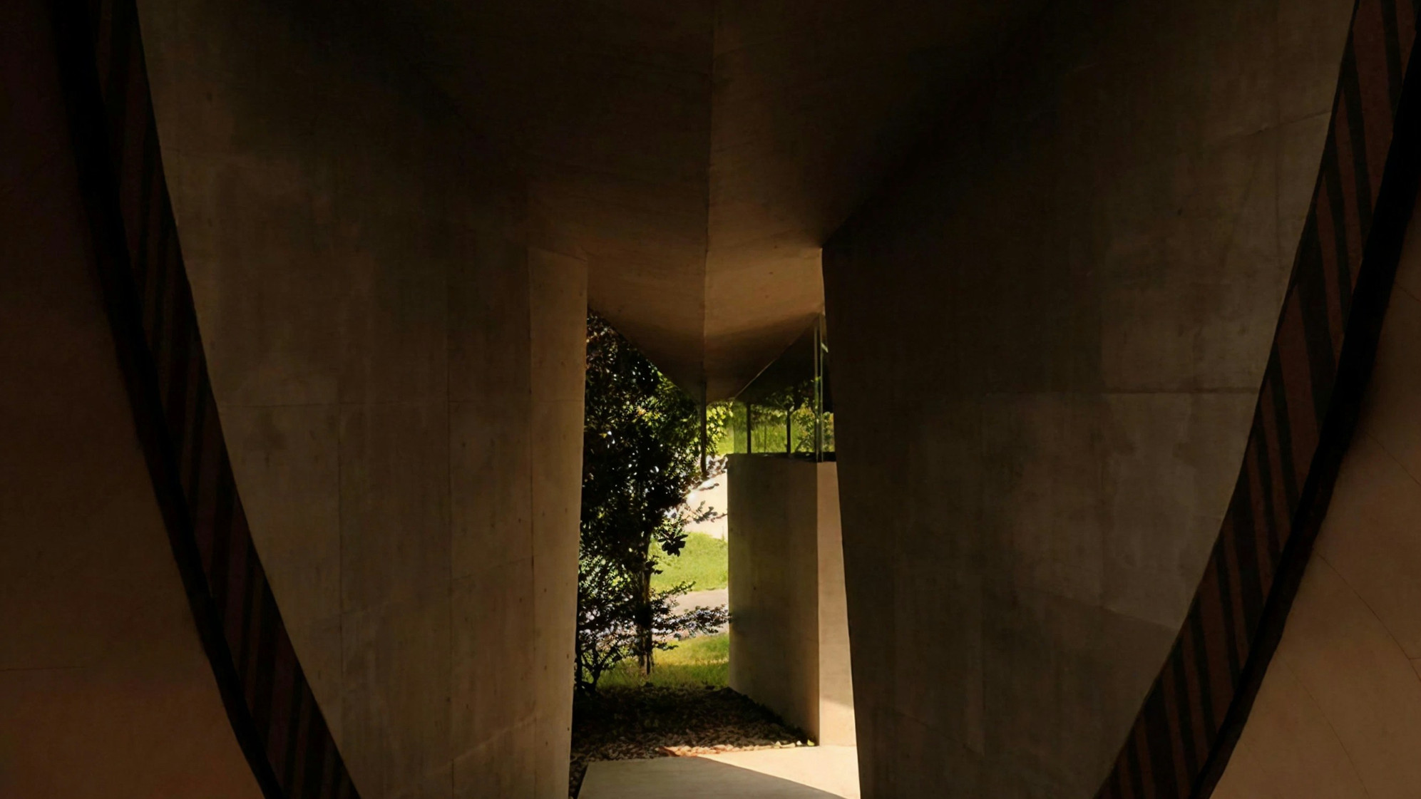Dim modern corridor with angular walls leading to a sunlit opening with trees and grass outside.
