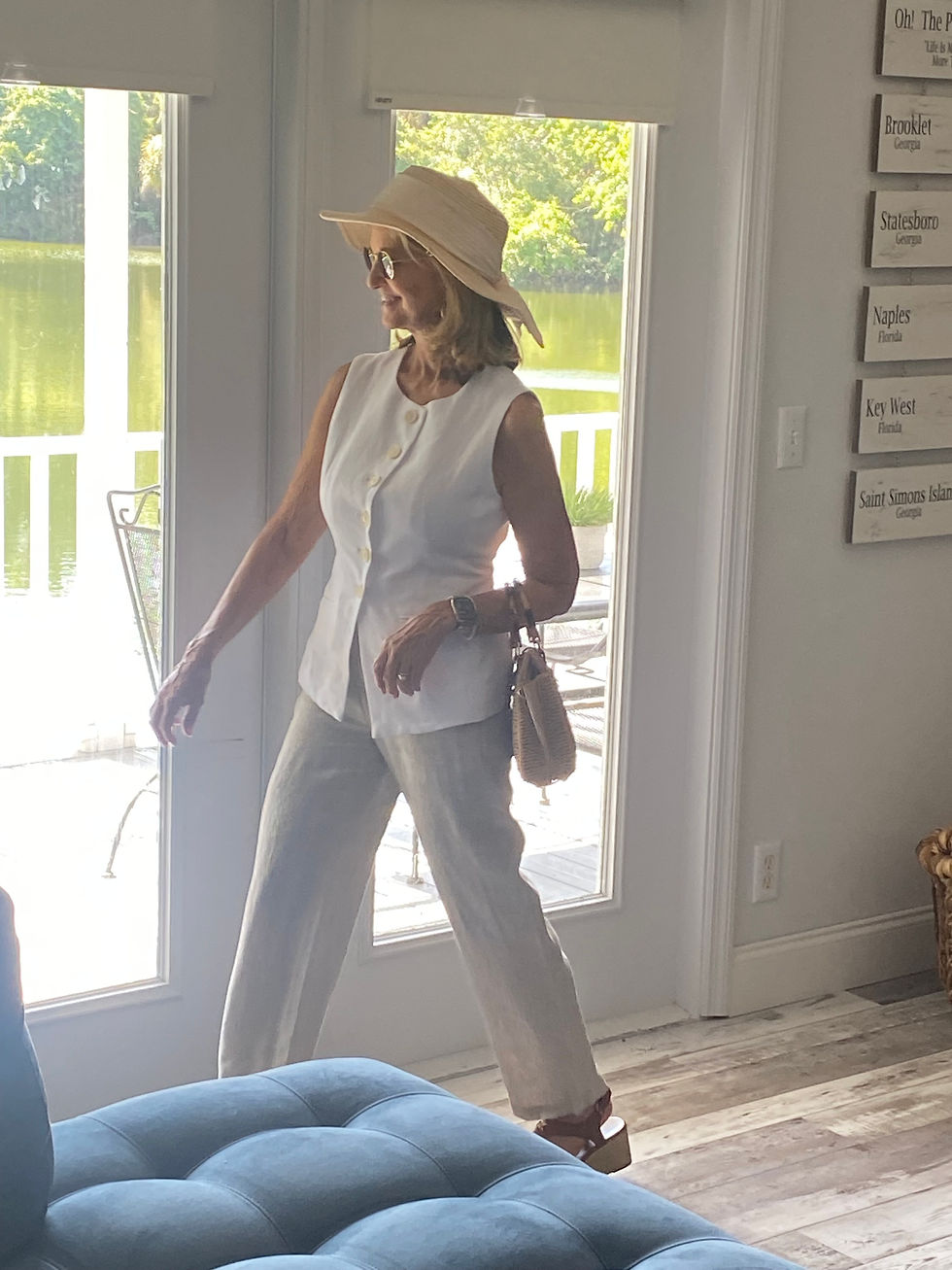 a petite woman walking out the door wearing a white vest, tan linen pants, and a cream colored sunhat