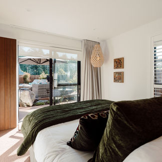 Cozy master ensuite bedroom with forest green bed covers, wood accents, and a Koura David Trubridge light with wall art by NZ artist Helen Keene. Glass door reveals a sunlit patio with garden view. Relaxing mood.