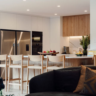 Modern chefs kitchen with wooden cabinets, stainless steel fridge, and a marble counter. Five wooden bar stools. Fruits and books add color.