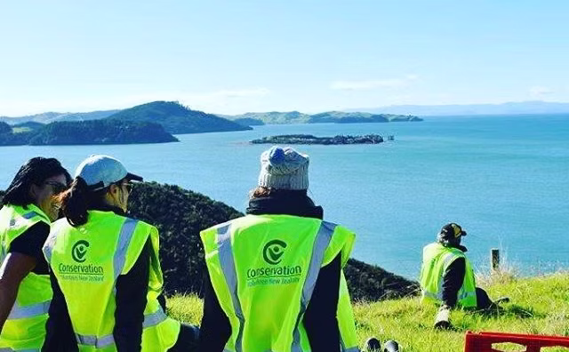 Four people in bright yellow high-visibility vests on the coast, with text promoting Conservation. Looking out towards a coastline.
