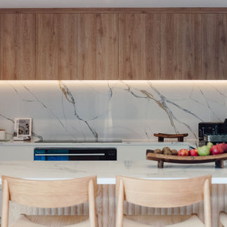 Modern kitchen with marble backsplash, wooden cabinets, and white countertops. Fruit bowl and kitchen items arranged neatly. Warm ambiance.