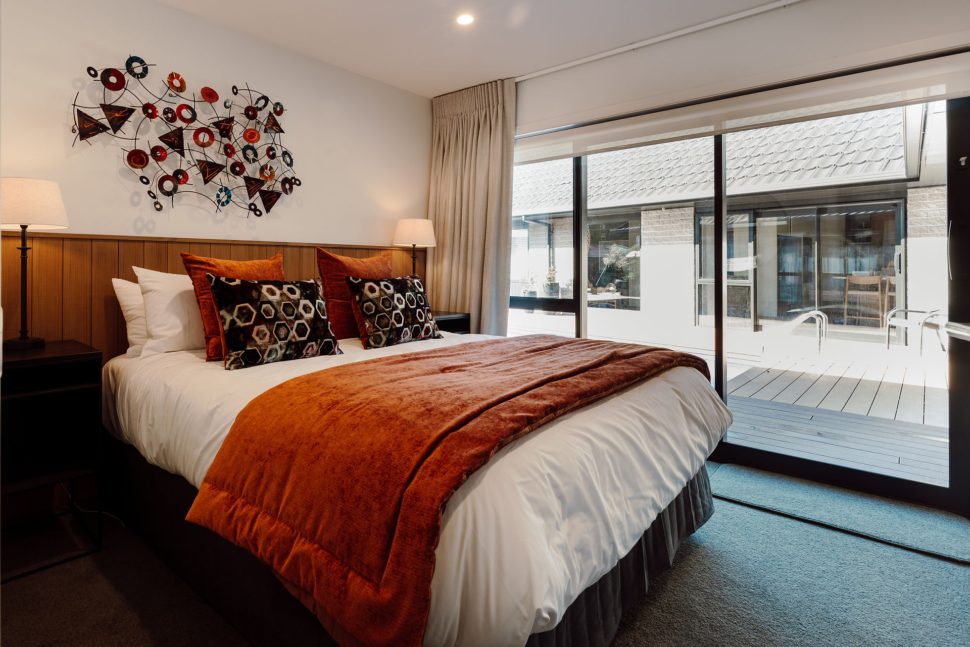 Modern bedroom with a and copper toned bedspread, decorative hexagon pillows, metal wall art, and large windows overlooking swimming pool. Warm, cozy ambiance.