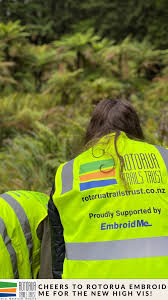 Two people in bright yellow high-visibility vests in a forest, with text promoting Rotorua Trails Trust and Embroid Me. Lush greenery surrounds them.