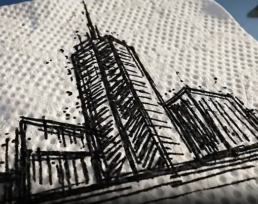 A close-up, angled shot of a black ink sketch of a city skyline, featuring a prominent skyscraper with a tall spire, drawn on a textured white paper towel. The drawing style is gritty and expressive, with visible ink splatters and hatched lines. The background is slightly blurred with a soft blue sky and a dark object in the upper right corner.