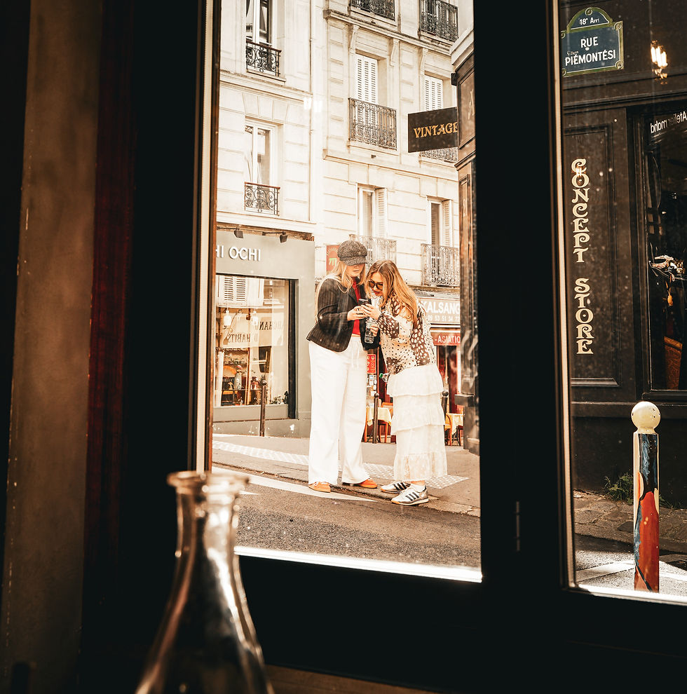 Two women in stylish outfits look at a phone together on a city street. They're viewed through a café window, with a sign reading "CONCEPT STORE."