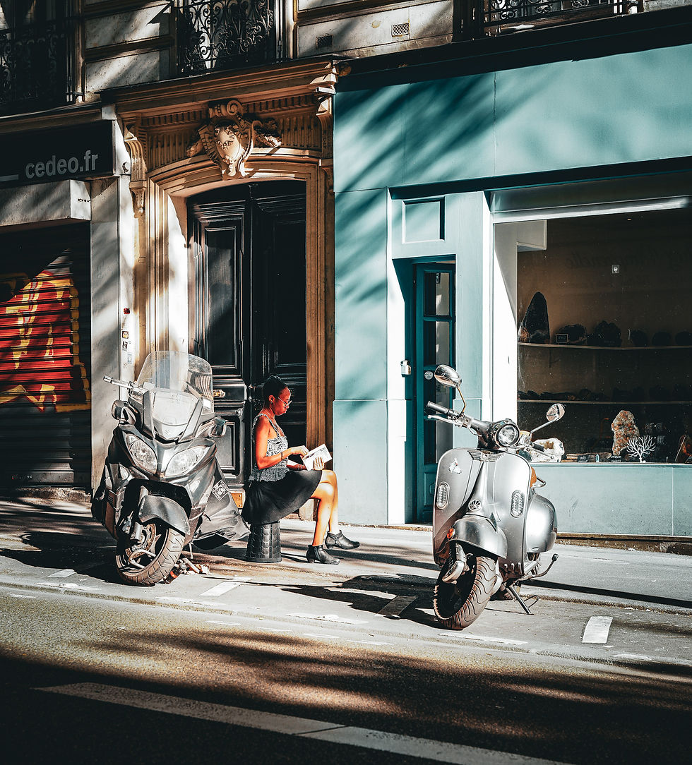 Woman sitting on a stool reads a magazine between two parked scooters on a sunlit street. Blue building facade in the background.