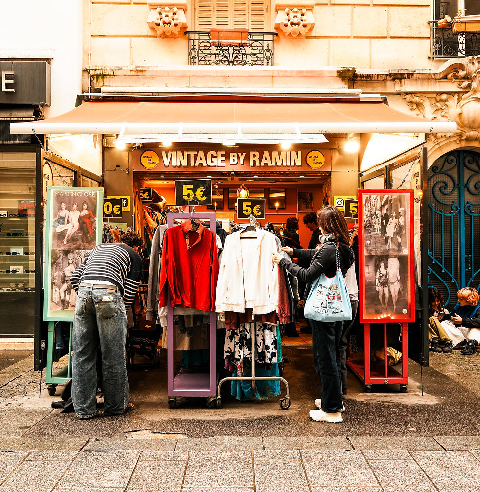 People browsing clothes at a vintage store with a sign "VINTAGE BY RAMIN." Bright colors and 5€ tags on racks. Parisian street vibe.