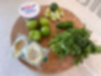 Ingredients on wooden board: sour cream, tomatillos, lime wedges, jalapeño, cilantro, and frozen tortillas. Bright kitchen setting.