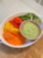 A white bowl with sliced bell peppers, carrots, broccoli, and snap peas, alongside a small bowl of green dip on a textured surface.