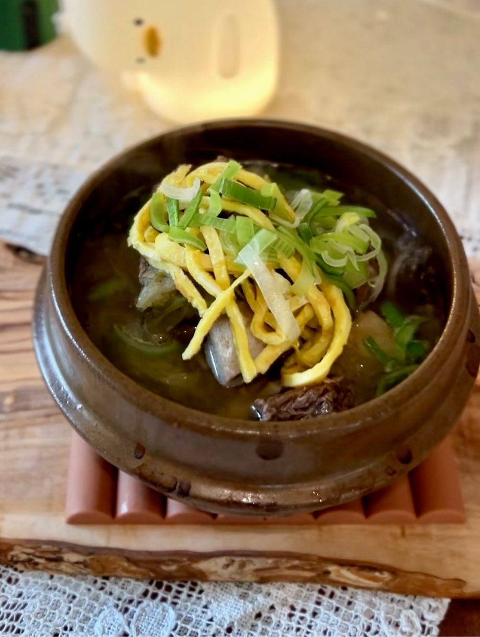 A ceramic bowl of soup with sliced egg and scallions sits on a wooden mat, lace tablecloth underneath, warm ambiance with a yellow light.
