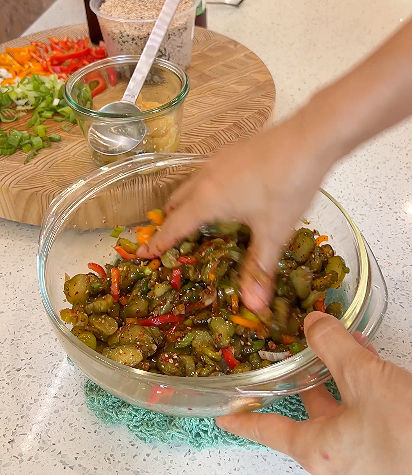 투명한 유리그릇에 피클을 손으로 버무리는 모습. 배경에는 채소가 있는 도마. 조리하는 활동감 있는 분위기.