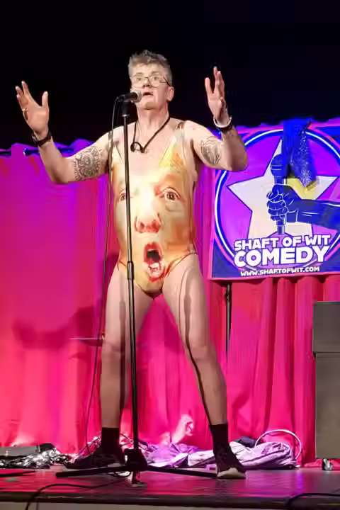 Joe Pasquale in a Donald Trump Leotard stands on stage with his arms raised, engaging the audience with enthusiasm.