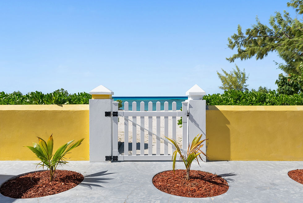 White picket gate opens to a sandy beach under a clear blue sky. Yellow wall and green plants frame the scene, creating a tranquil vibe.