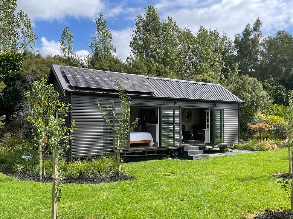 A dark grey Tiny House with its doors open, sits in a lush green yard, with a nice lawn out the front of it and trees behind.