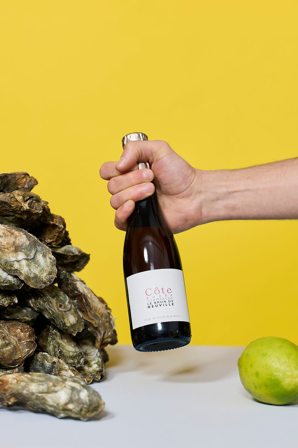 Hand hält eine Flasche Le Brun de Neuville Champagne Côte Rosée neben frischen Austern und Limette vor gelbem Hintergrund.