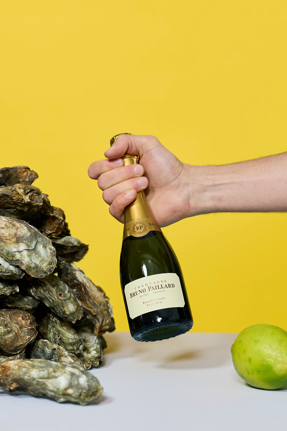 Hand hält eine Flasche Bruno Paillard Champagne Premier Cuvée neben frischen Austern und Limette vor gelbem Hintergrund.