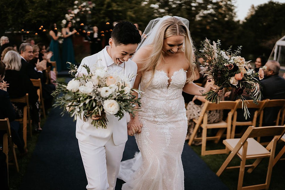 Newlywed couple in wedding attire walk hand-in-hand down an outdoor aisle, holding bouquets.