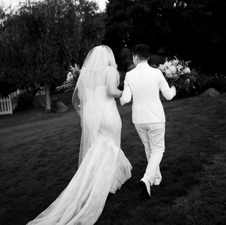 A black and white photo of a newlywed couple walking away on a grassy lawn.