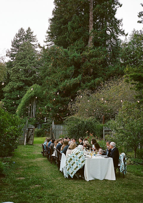 Guests seated at a long dining table outdoors, surrounded by lush trees and string lights. The setting conveys a warm, intimate gathering.