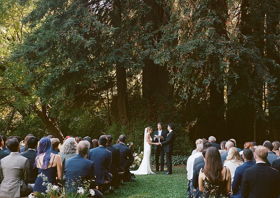 Outdoor wedding ceremony at Dawn Ranch with guests seated on wooden benches. The bride and groom stand beneath tall trees on a grassy area, flanked by floral arrangements.