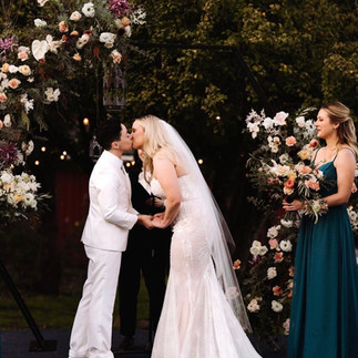 A couple kissing under a floral arch at an outdoor wedding ceremony.