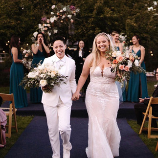 Two brides smiling and walking down an aisle, each holding a bouquet.