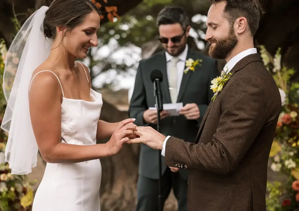 Bride in a white dress and groom in a brown suit exchanging rings at an outdoor wedding ceremony.