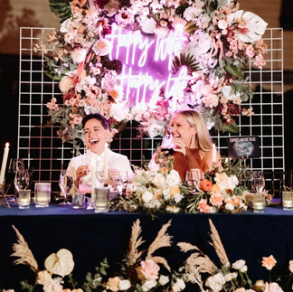 Two people seated at a decorated table with a pink neon sign reading "Happy Wife Happy Life" amid floral arrangements.
