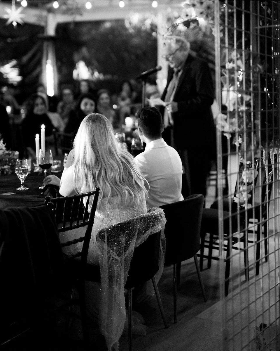 A black and white image of a wedding with a speaker at a microphone and attendees seated at tables.