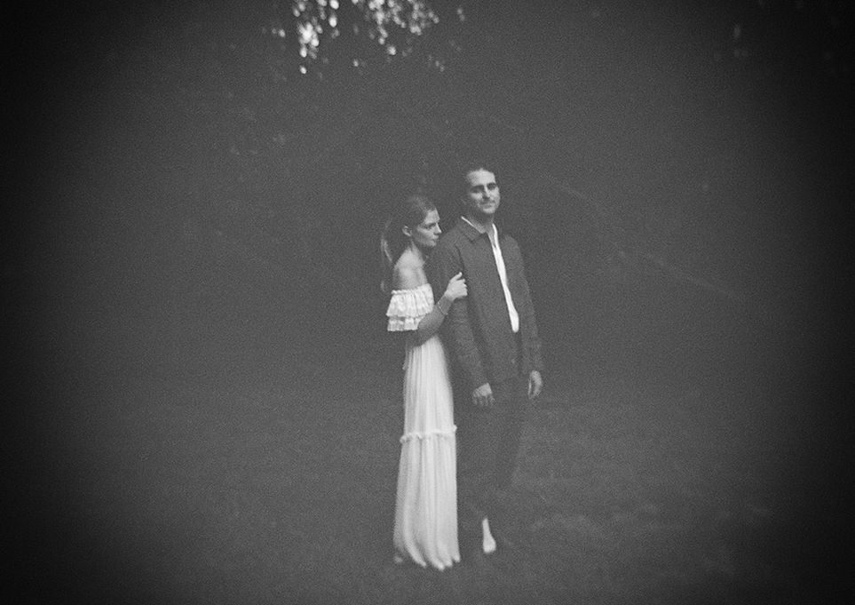 Two people stand closely in a dreamy, black-and-white photo. The woman, in an off-shoulder dress, rests her head on the man’s shoulder; both appear serene.