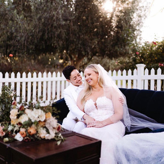 A happy couple sitting on a dark blue couch outdoors, with a bouquet of flowers in the foreground and a white picket fence in the background.