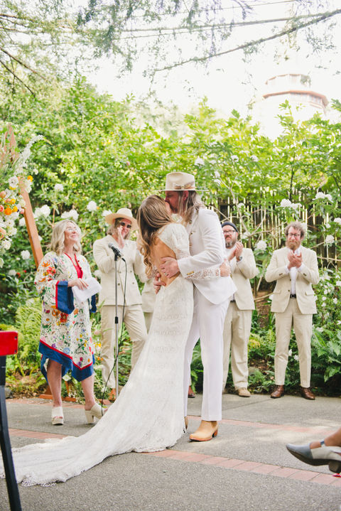 A bride and groom share a kiss at an outdoor wedding ceremony. The bride wears a long lace gown, and the groom is in a white suit with a hat. Guests applaud.