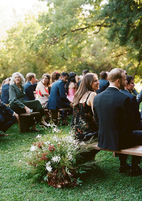 Guests in formal attire sit on wooden benches at an outdoor wedding, surrounded by lush greenery and flowers. The scene is sunny and joyful.