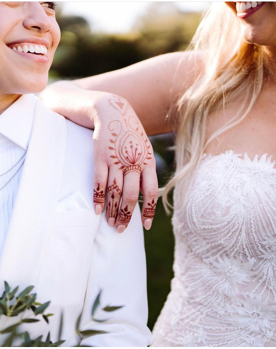 Two people in wedding attire, one with henna on their hand resting on the other's shoulder.